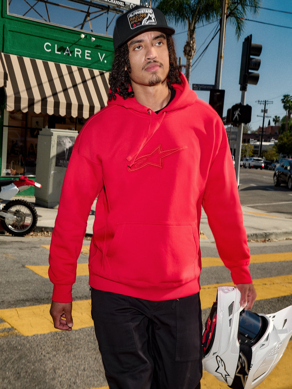 Alpinestars Emergent Oversized hættetrøje, rød, 3D-broderet Astars-logo på brystet, kombineret med sorte bukser og en kasket, mens han bærer en hvid motocross-hjelm, afslappet urban streetwear-stil