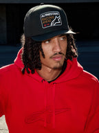 Mand iført Alpinestars Intuitive Snapback-kasket i sort, med struktureret krone og 'Racing Ahead' broderet logo-patch, kombineret med en klar rød hættetrøje, afslappet lifestyle streetwear