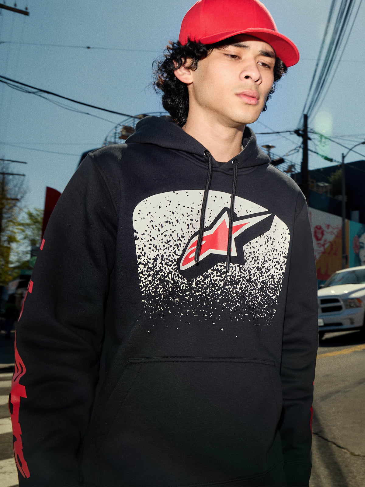 Alpinestars Obsessed hættetrøje, sort pullover-fleece, hvid MX-graduering grafik på brystet med rødt stjernelogo, røde wordmark-logoer på ærmerne, afslappet motorsport-inspireret streetwear i et urbant miljø.