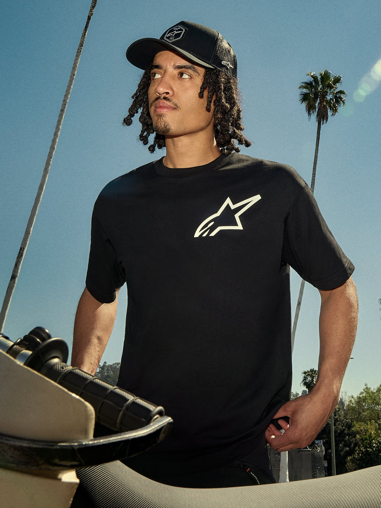Alpinestars One Oversized CSF Tee, kortærmet T-shirt, sort med hvidt Astars-logo, bæres af en mand i en sort trucker-hat, udendørs livsstilsmiljø med palmer og en motorcykel, afslappet motorsport-streetwear