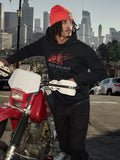 Alpinestars Pulse Hoodie, sort hættetrøje i fleece med rødt og gråt Astars Combo-logo, båret af en mand med en orange hue, der står ved siden af en rød Honda-motorcykel i et bymiljø, casual motorsport-streetwear