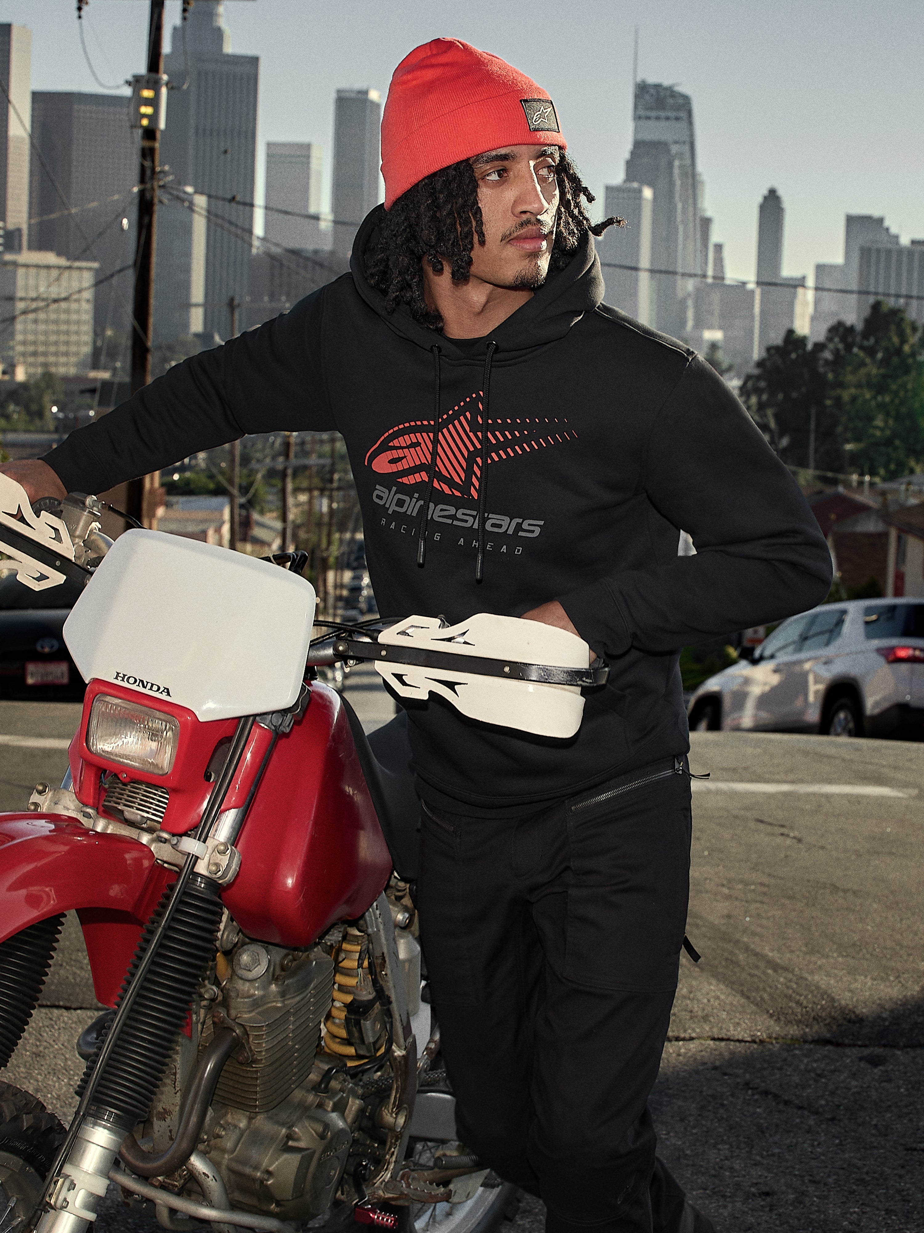 Alpinestars Pulse Hoodie, sort hættetrøje i fleece med rødt og gråt Astars Combo-logo, båret af en mand med en orange hue, der står ved siden af en rød Honda-motorcykel i et bymiljø, casual motorsport-streetwear