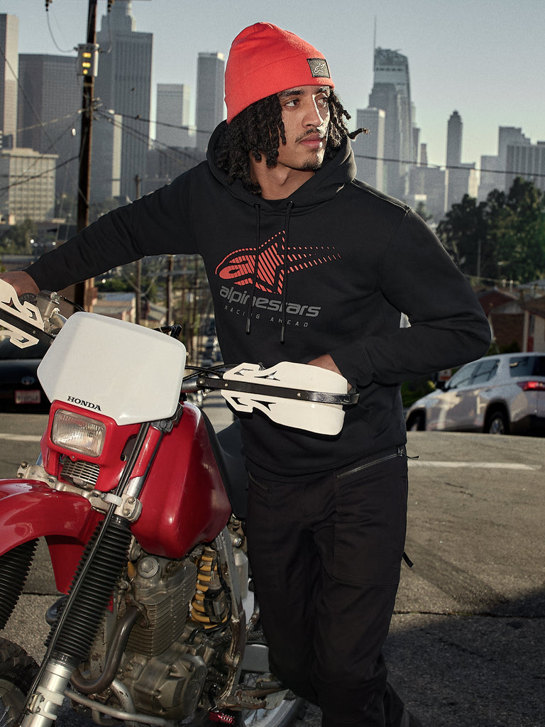 Alpinestars Pulse Hoodie, sort hættetrøje i fleece med rødt og gråt Astars Combo-logo, båret af en mand med en orange hue, der står ved siden af en rød Honda-motorcykel i et bymiljø, casual motorsport-streetwear