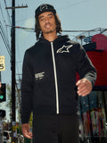 Alpinestars Transverse Hoodie, sort fleece med lynlås foran, hvid lynlås og hvidt Astars-logo på brystet, 'One Goal One Vision' tekstprint, afslappet streetwear-stil, båret af en mand i et urbant udendørsmiljø