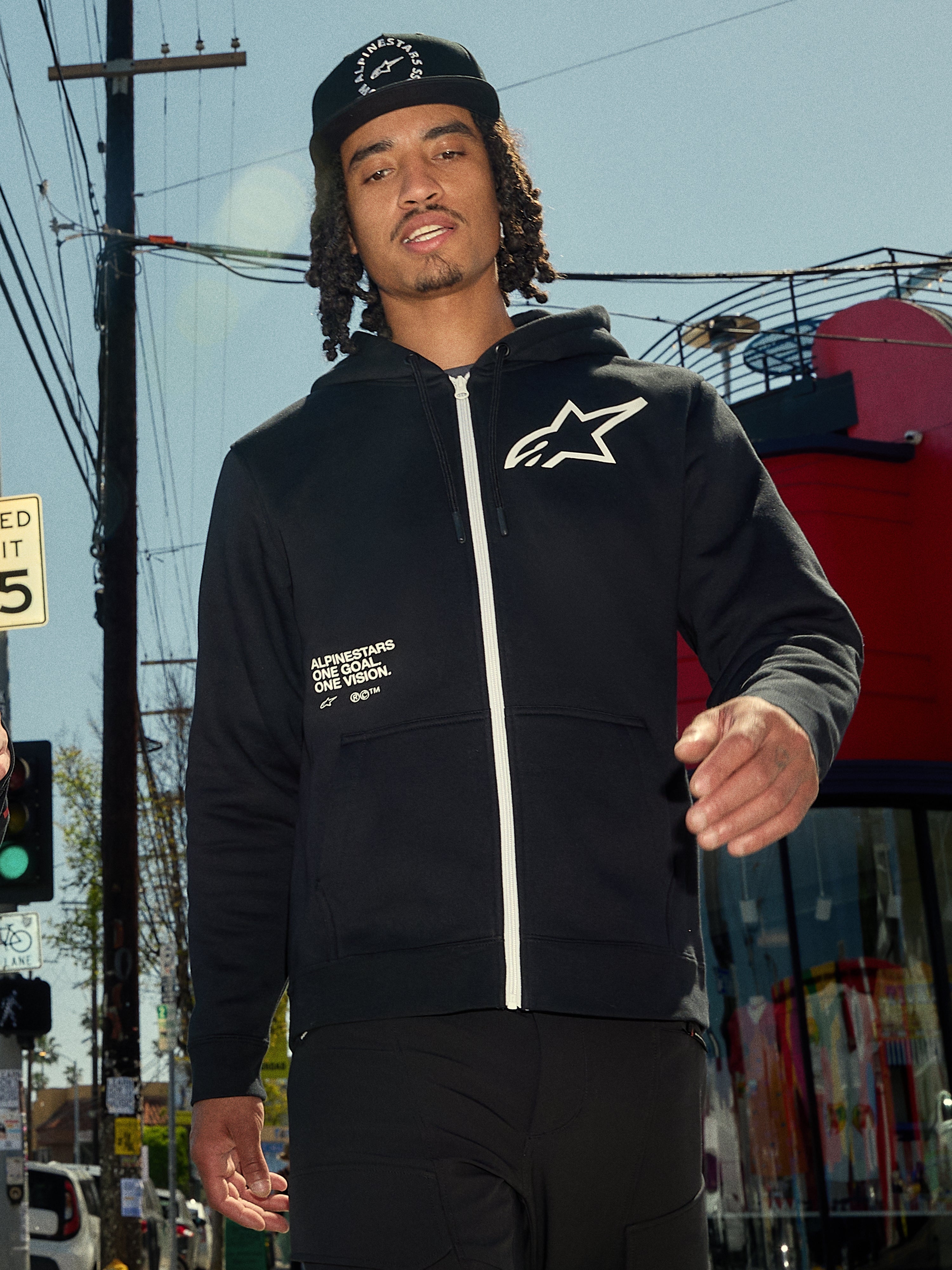Alpinestars Transverse Hoodie, sort fleece med lynlås foran, hvid lynlås og hvidt Astars-logo på brystet, 'One Goal One Vision' tekstprint, afslappet streetwear-stil, båret af en mand i et urbant udendørsmiljø