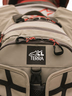 Alpinestars AT-15 rygsæk, Aluminum Walnut, nærbillede af All Terra-logomærke på sandfarvet teknisk tekstil, med orange lynlåsstropper og robuste konstruktionsdetaljer