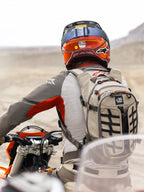 Alpinestars AT-15 rygsæk i Aluminum Walnut, båret af en adventure-motorcykelrytter i et ørkenlandskab, robust beige tekstilkonstruktion med sorte webbing-stropper, orange lynlåsdetaljer og 'All Terra'-branding, kombineret med en rød og orange hjelm og gråt køreudstyr til off-road touring.