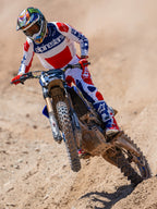 Motocrosskører i aktion iført Alpinestars Limited Edition Fortitude Tech 10-støvler, klar rød, hvid og blå racing-trøje og bukser, matchende patriotisk udstyrssæt til off-road kørsel på en jordbane.