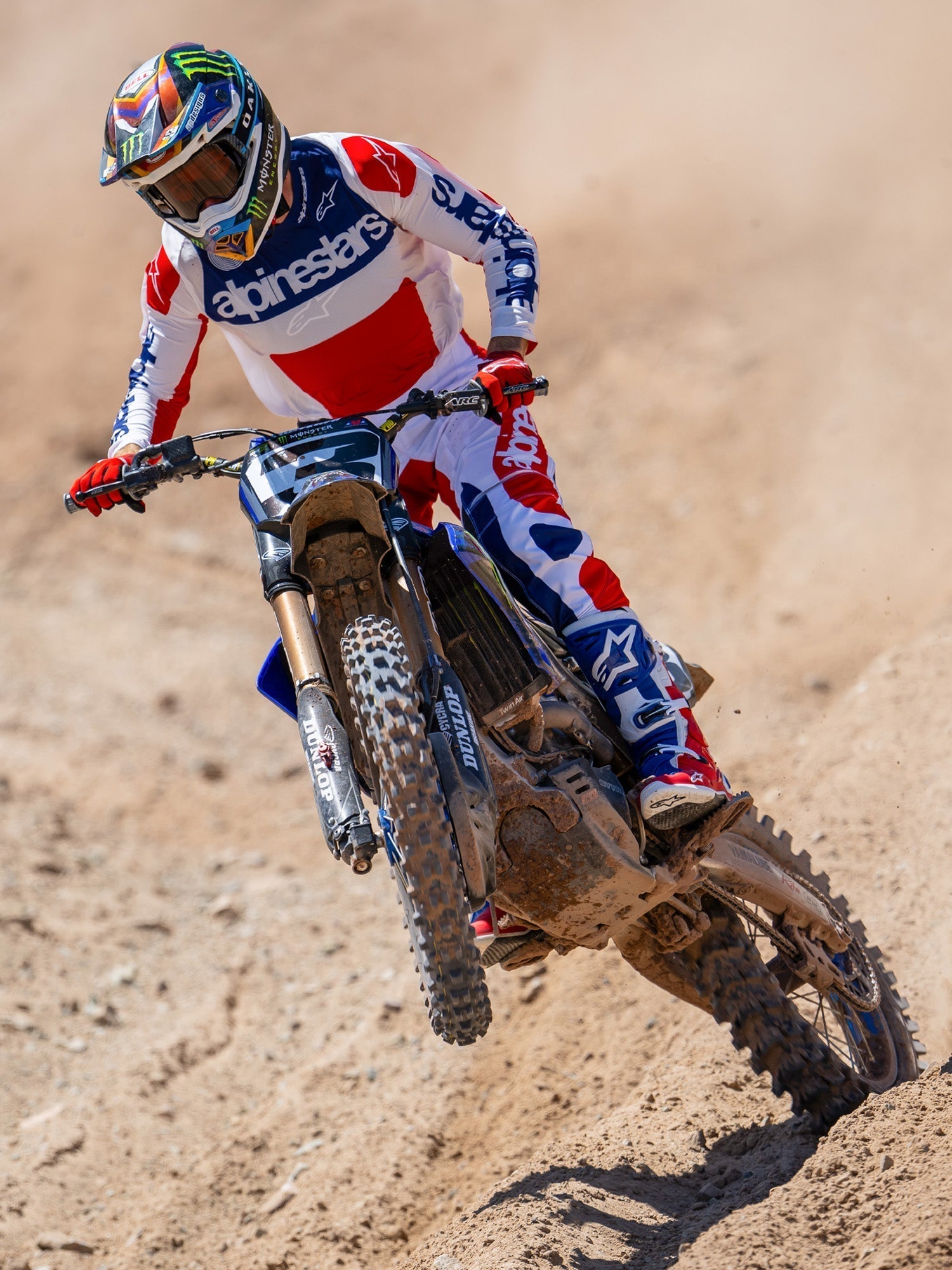 Motocrosskører i aktion iført Alpinestars Limited Edition Fortitude Tech 10-støvler, klar rød, hvid og blå racing-trøje og bukser, matchende patriotisk udstyrssæt til off-road kørsel på en jordbane.