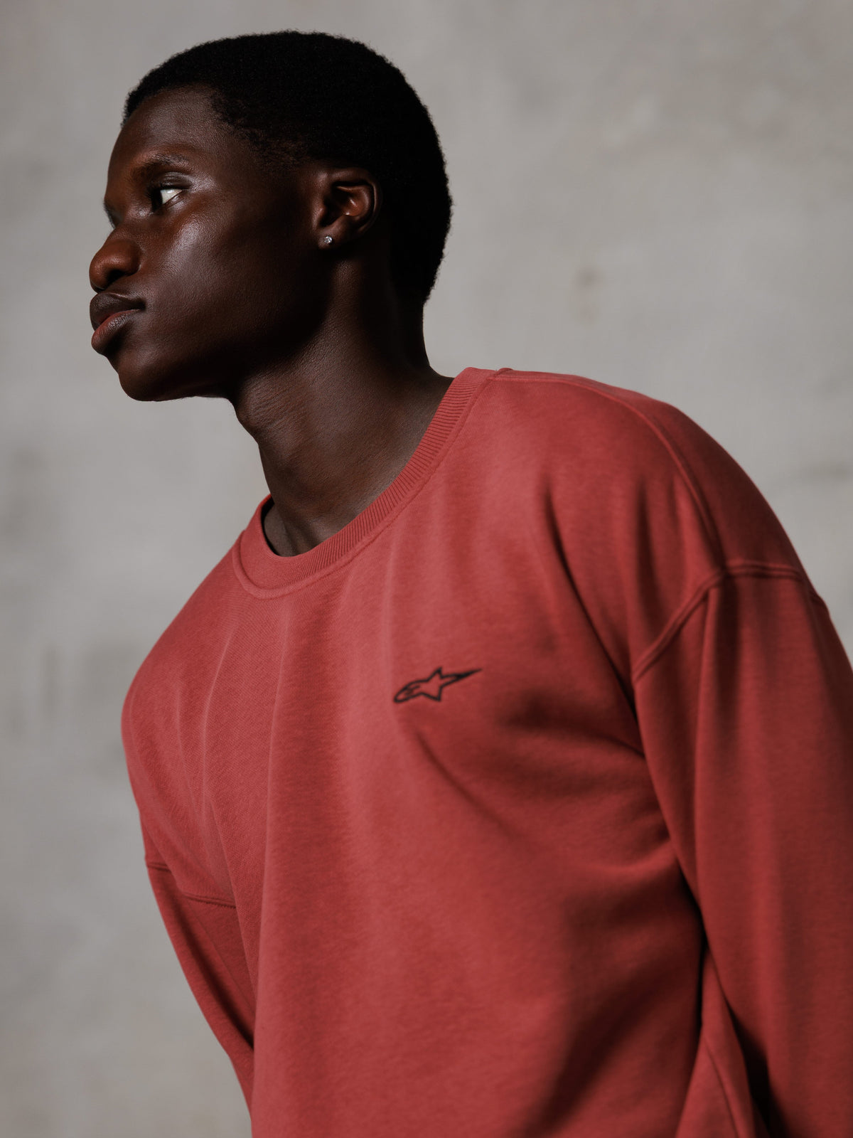 Alpinestars Essentials Ageless Crew, teglrød crewneck sweatshirt, båret af en mandlig model i sideprofil, lille sort stjernelogo på brystet, afslappet pasform, premium fleece, afslappet streetwear