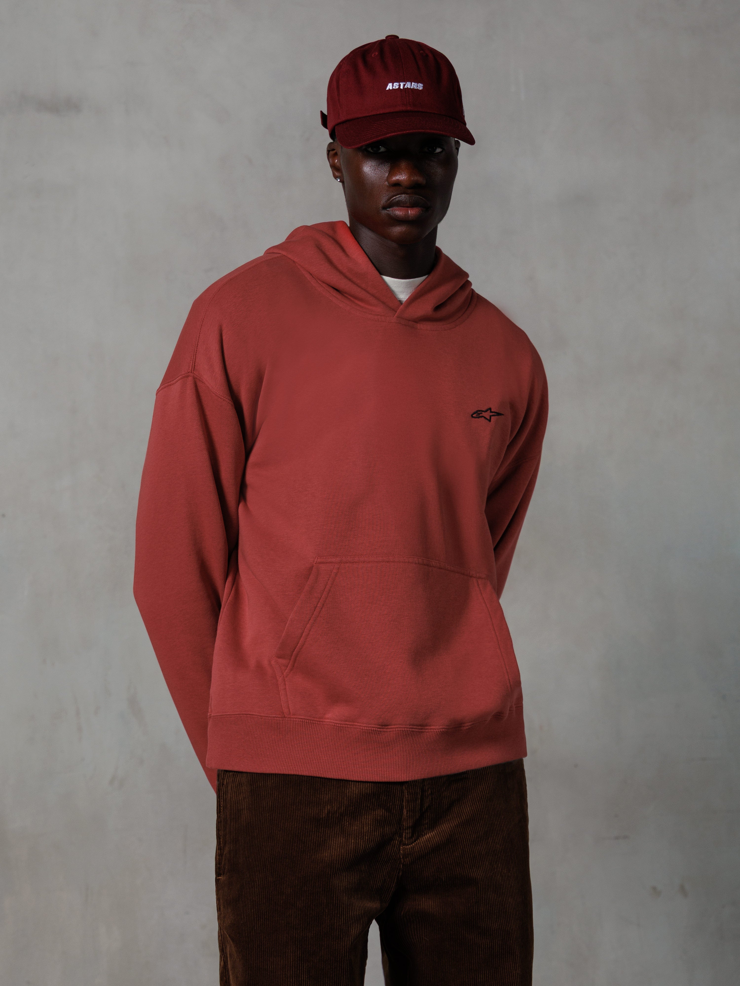 Alpinestars Essentials Ageless hættetrøje, afslappet streetwear, teglrød, lille sort brystlogo, kængurulomme foran, båret med en matchende rød ASTARS kasket og brune fløjlsbukser, rent minimalistisk design