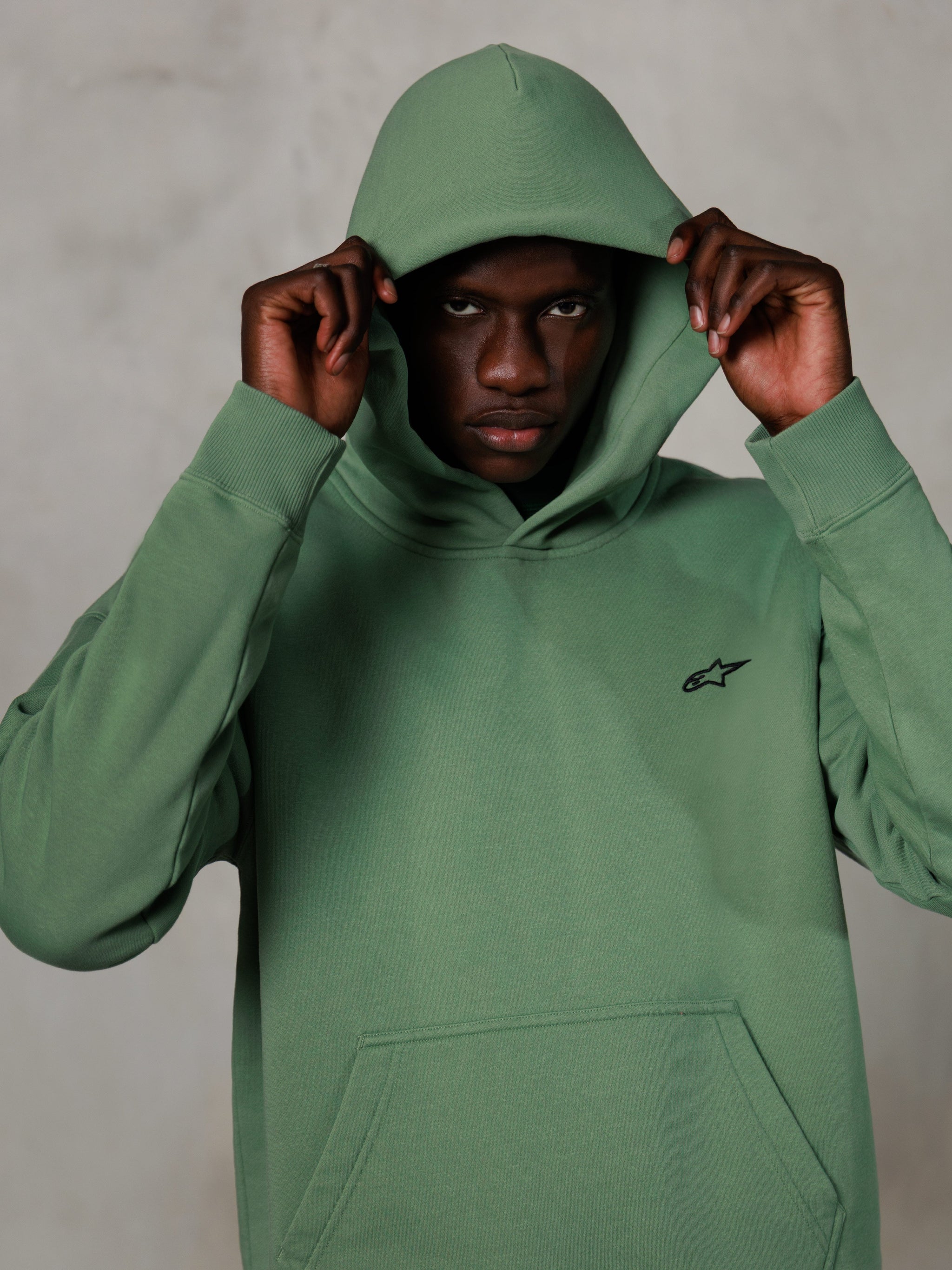Alpinestars Essentials Ageless Hoodie, casual streetwear, cactus green with small black logo on chest, model wearing hoodie with hood up, front pouch pocket, relaxed motorsport-inspired design