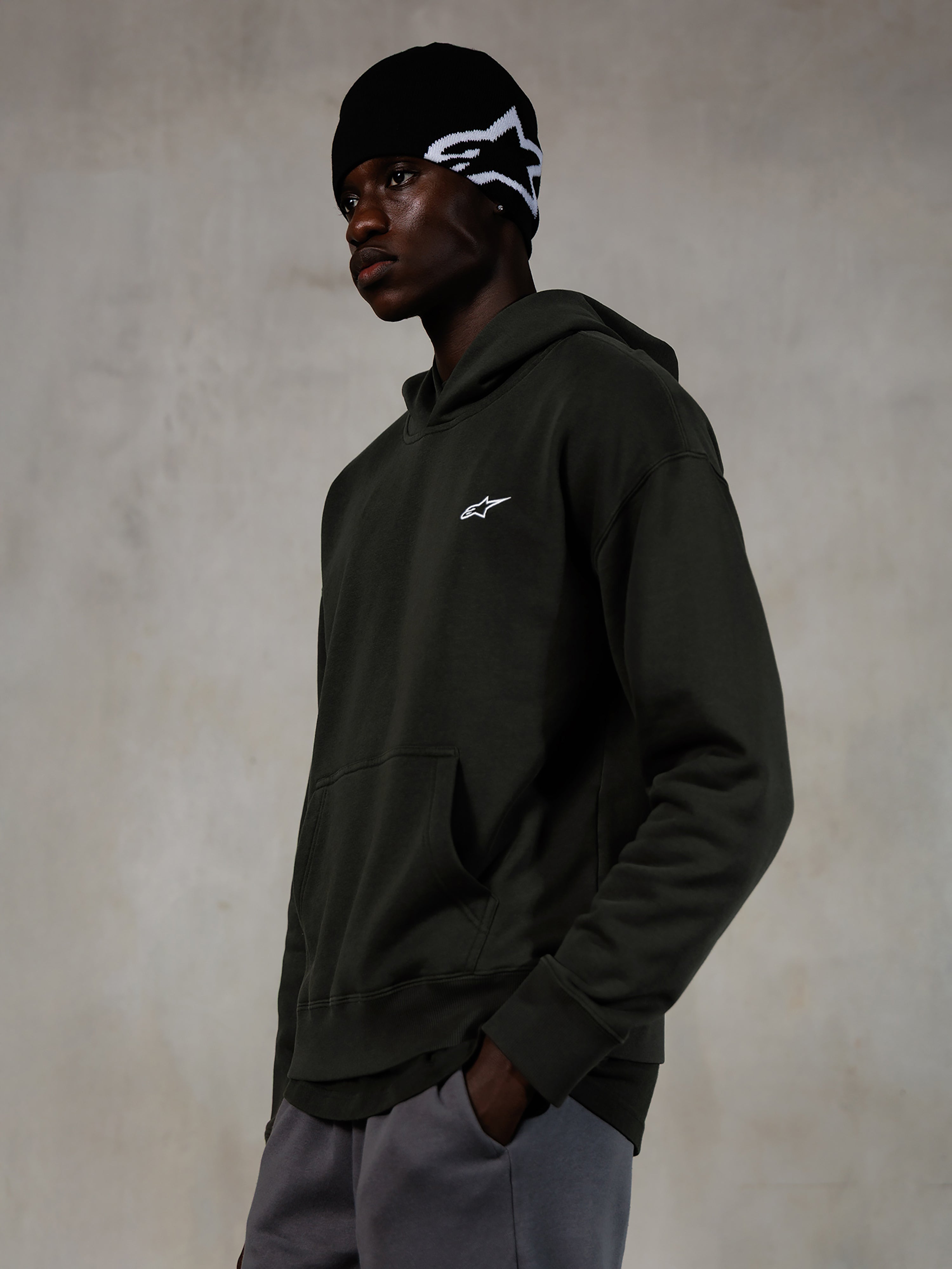Alpinestars Essentials Ageless Hoodie, Off Black, mandlig model iført afslappet streetwear, mørk koksgrå stenvasket fleece med et lille hvidt stjerne-logo, kombineret med en sort Alpinestars beanie og grå bukser