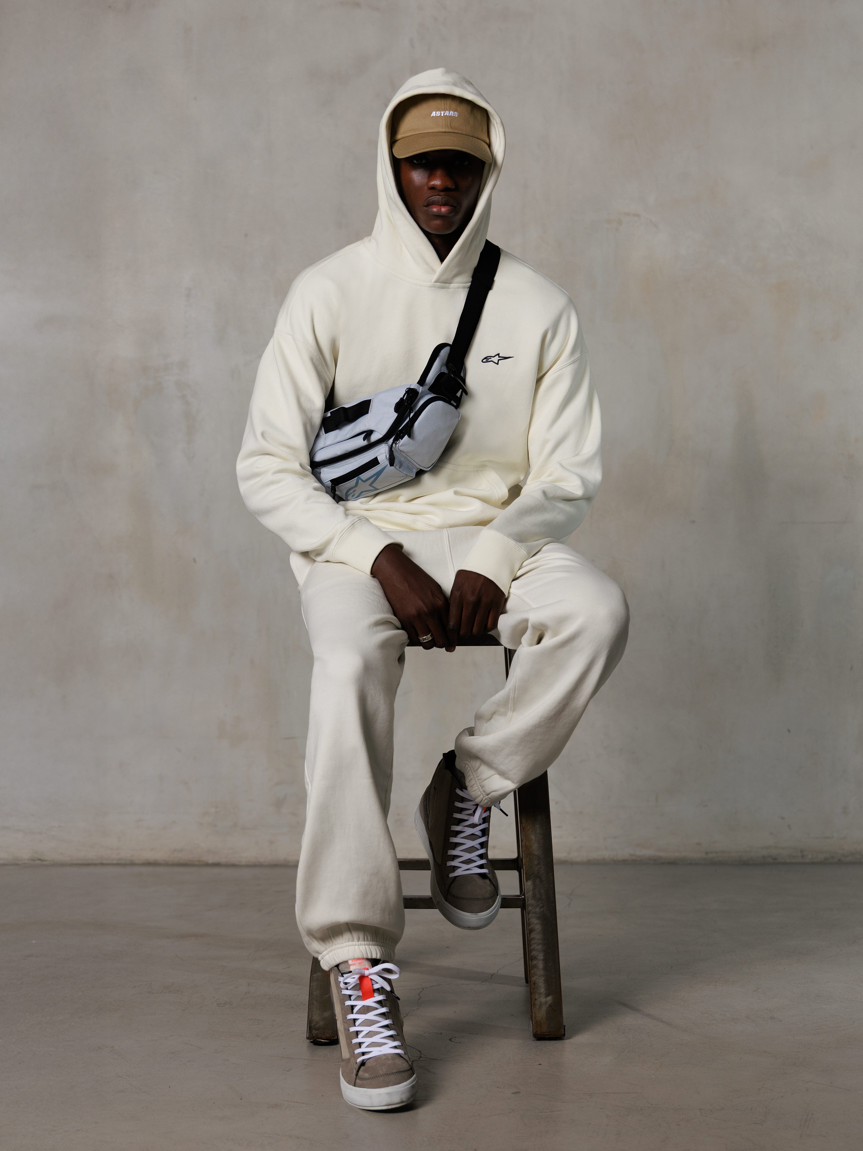 Alpinestars Essentials Ageless Hoodie, Off-White, båret af en model på en taburet med matchende fleece-joggers, sandfarvet ASTARS-kasket og crossbody-taske, afslappet motorsport-inspireret streetwear
