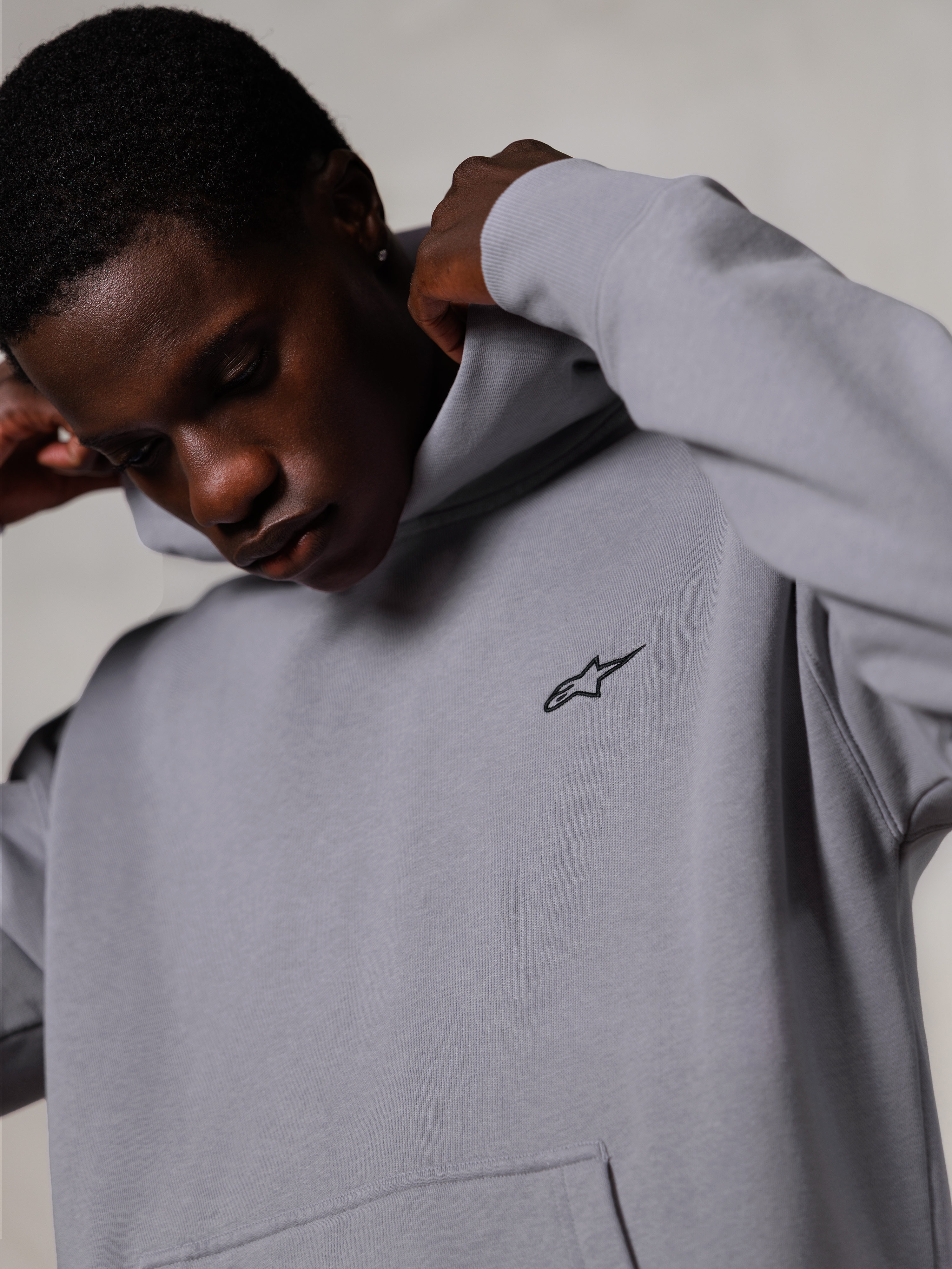 Alpinestars Essentials Ageless Hoodie, Silver, nærbillede af en mand, der justerer hætten på den grå hættetrøje, lille sort Alpinestars-logo på brystet, førsteklasses motorsportsinspireret fritidstøj