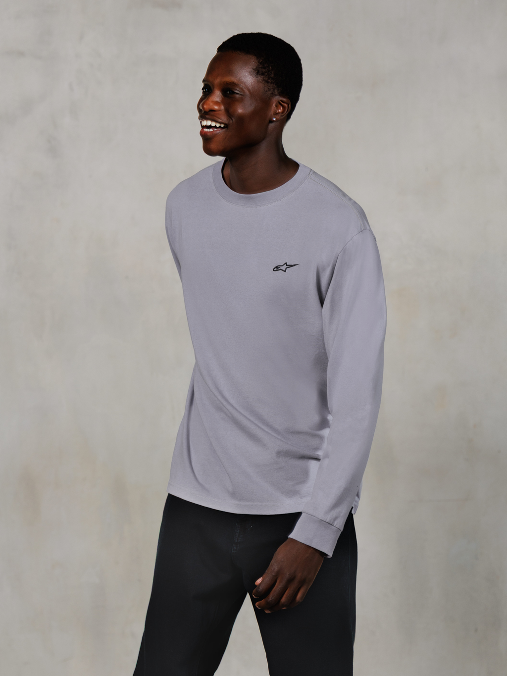 Alpinestars Essentials Ageless LS Tee, langærmet t-shirt, sølv, lille sort logo på brystet, båret af en smilende model med mørke bukser, afslappet moderne pasform, afslappet motorsport-livsstilstøj