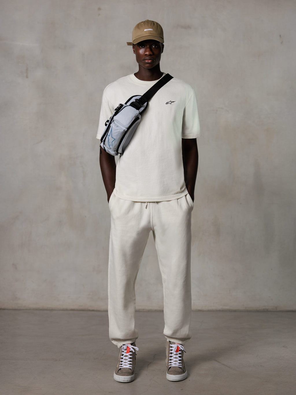 Alpinestars Essentials Ageless SS Tee, kortærmet T-shirt, Off White, med et lille sort logo på brystet, båret med matchende joggers, beige baseball cap og en grå crossbody-taske, afslappet lifestyle streetwear