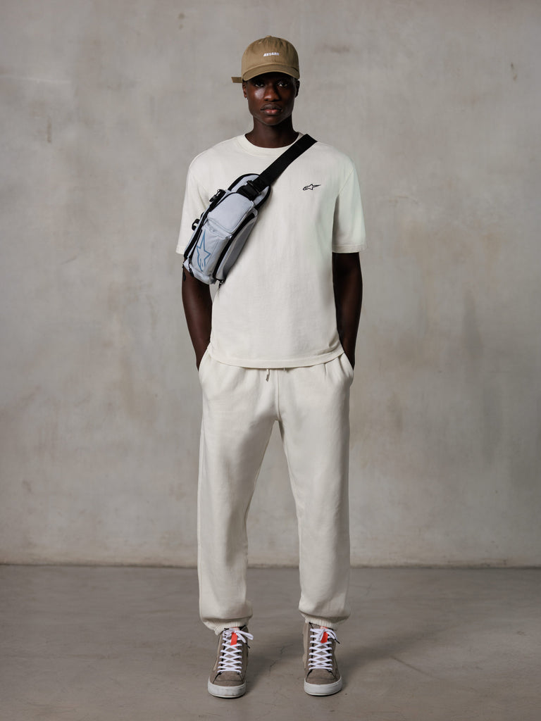 Alpinestars Essentials Ageless SS Tee, kortærmet T-shirt, Off White, med et lille sort logo på brystet, båret med matchende joggers, beige baseball cap og en grå crossbody-taske, afslappet lifestyle streetwear