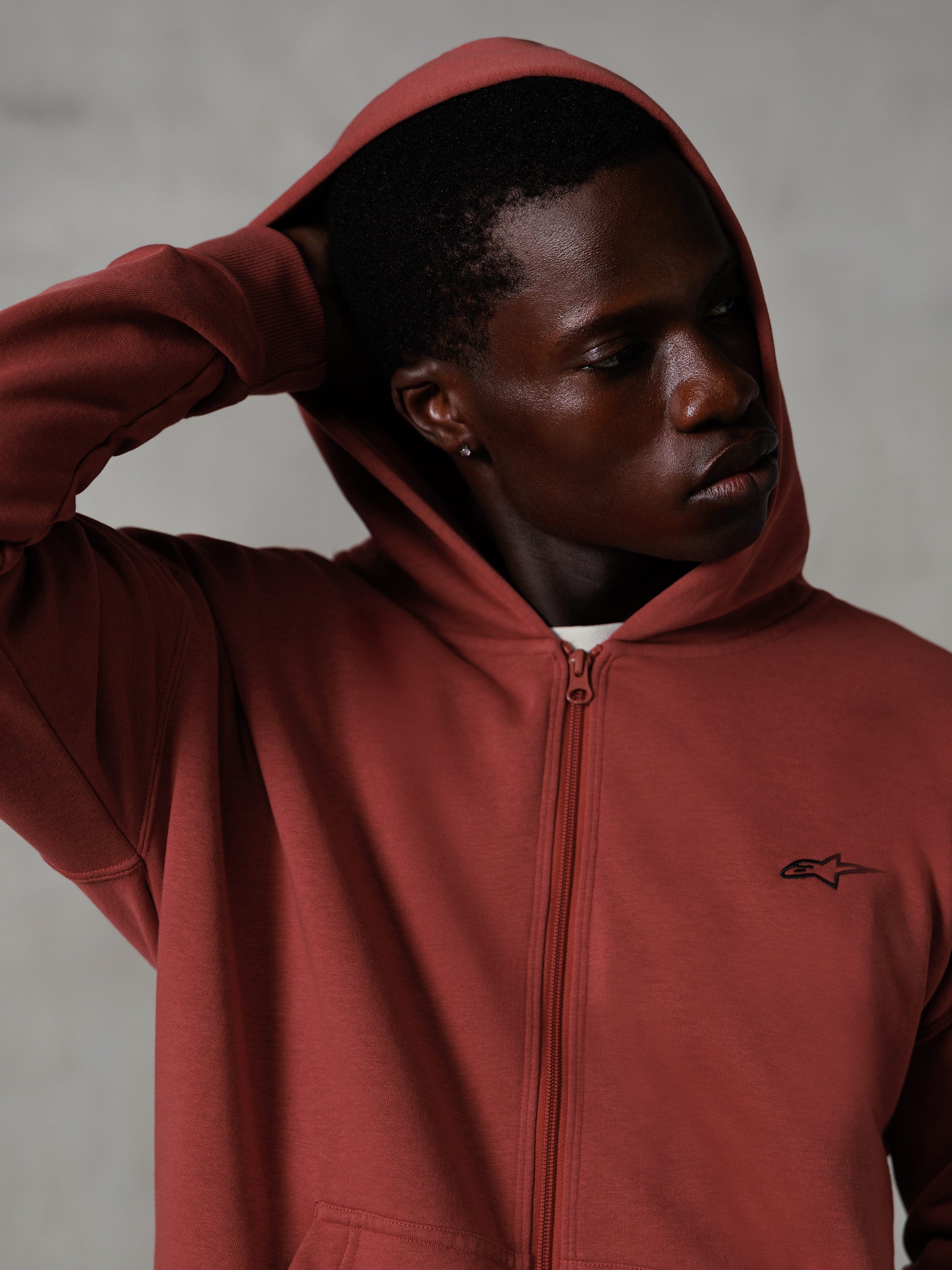 Alpinestars Essentials Ageless Zip Hættetrøje, teglrød, afslappet lifestyle streetwear, hætte oppe med hånd bag hovedet, lille sort Astars-logo på brystet, fleecehættetrøje med afslappet pasform