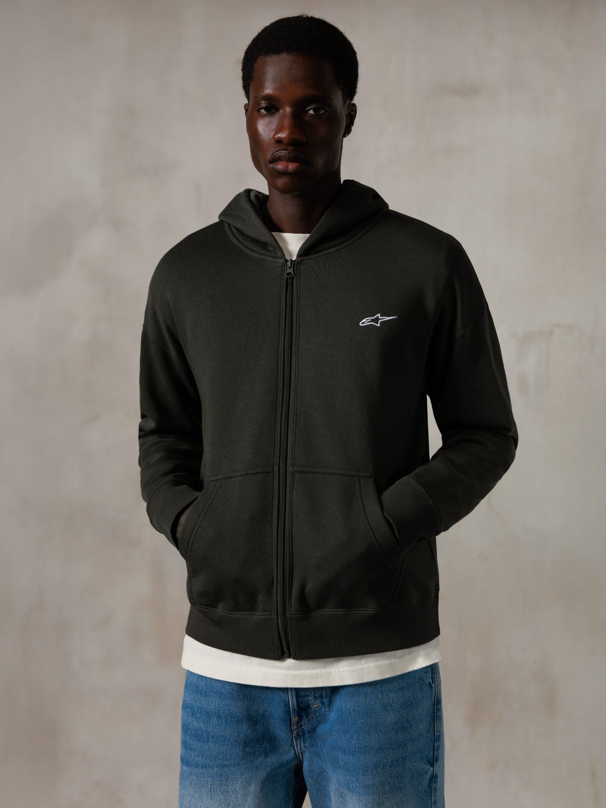Alpinestars Essentials Ageless Zip Hoodie, Off Black, hættetrøje med gennemgående lynlås og et lille hvidt Alpinestars-logo på brystet, afslappet pasform, båret af en model med blå jeans og hvid t-shirt, afslappet motorsport-inspireret streetwear