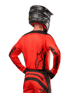 Motocrossrytter set bagfra iført Alpinestars 2024 Fluid Lurv trøje i Hot Orange og sort, med markant Alpinestars-branding på ryggen og ærmerne, kombineret med matchende motocrossbukser og en sort hjelm, designet til off-road racing.