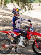Motocross rider wearing the Limited Edition HUNTA96 XXV Supertech Vision Goggles in Purple White Mirror Silver, matching purple and white racing jersey with white boots, seated on a red Honda dirt bike with number 96, outdoor off-road setting on a sandy track.