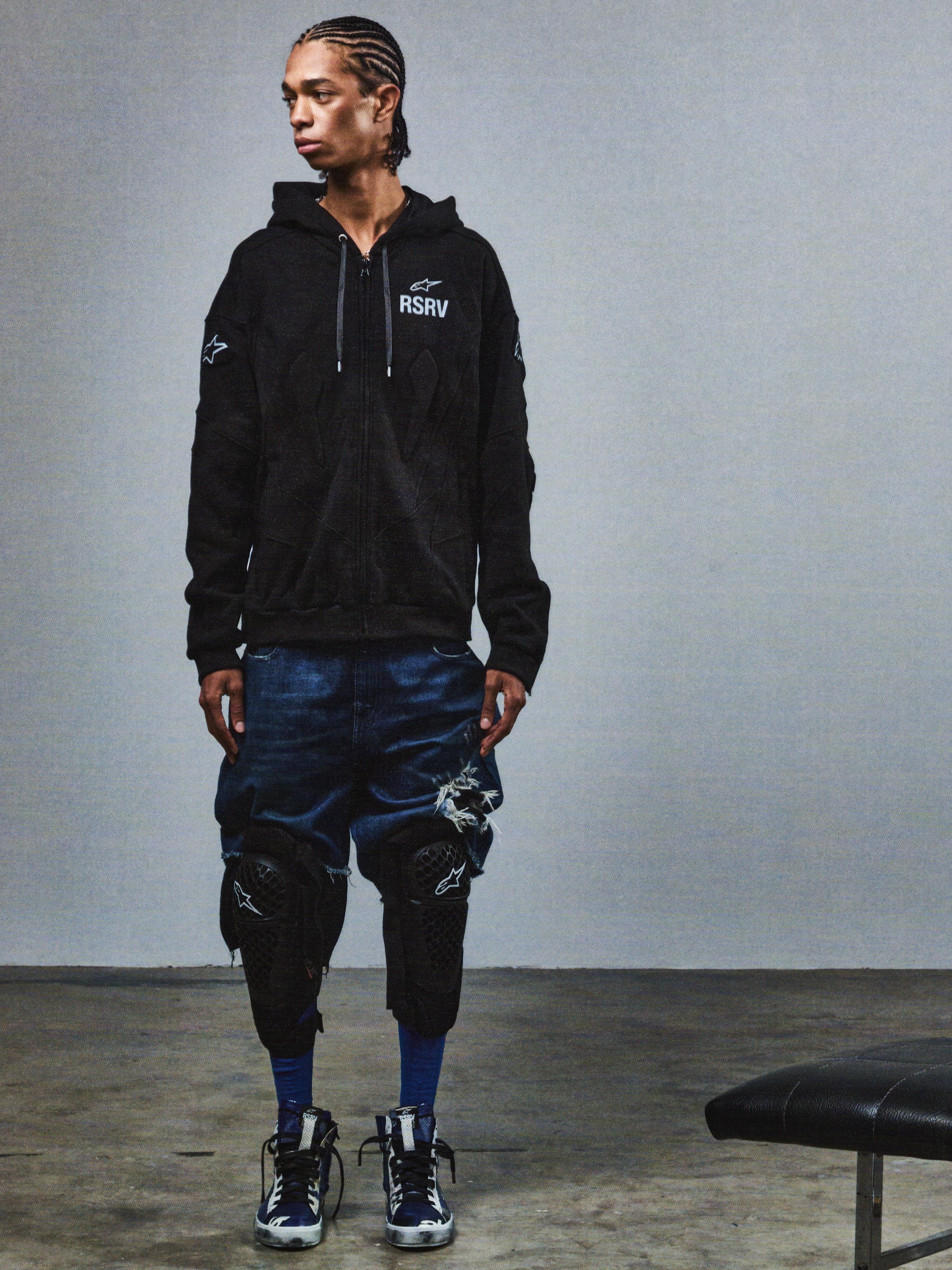 Alpinestars RSRV GP Hoodie, sort hættetrøje med RSRV-logo og stjerner, streetwear-kollektion, vist med distressed denimshorts og knæbeskyttere, premium motorsport-inspireret lifestyle-beklædning