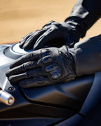 Alpinestars SP X 7 handsker, sorte motorcykelhandsker med kort manchet i perforeret læder, hænder hvilende på en mørk motorcykel-tank, med synlig TPU-knobeskyttelse med dobbelt densitet og præget branding, designet til sports- og landevejskørsel