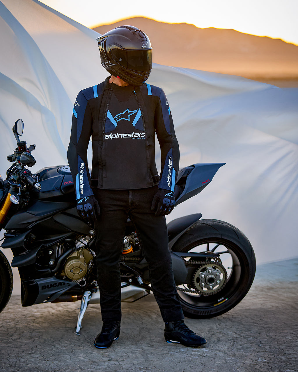 Alpinestars T-Stunt Air jakke, tekstiljakke med sportssnit, sort, mørkeblå og klar blå, båret af en motorcyklist ved siden af en Ducati-motorcykel, kulfiberhjelm og sort køreudstyr, i et ørkenlandskab ved solnedgang.
