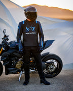 Alpinestars T-Stunt Air jakke, tekstiljakke med sportssnit, sort, mørkeblå og klar blå, båret af en motorcyklist ved siden af en Ducati-motorcykel, kulfiberhjelm og sort køreudstyr, i et ørkenlandskab ved solnedgang.