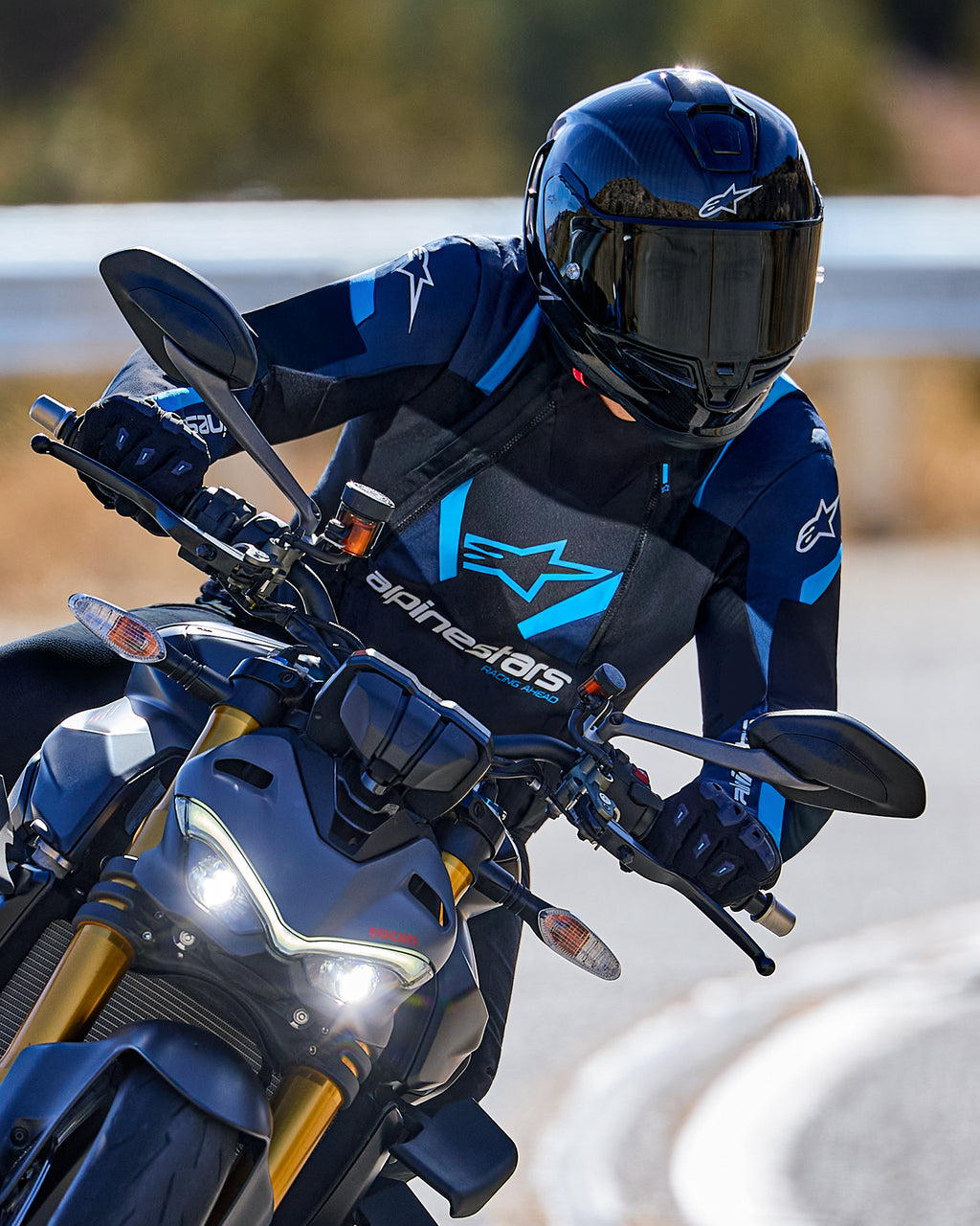 Motorcykelrytter der læner sig ind i et sving iført Alpinestars T-Stunt Air Jakke i sort, mørkeblå og lyseblå, kombineret med en sort hjelm og handsker, actionbillede på en asfalteret vej, sportsudstyr til kørsel