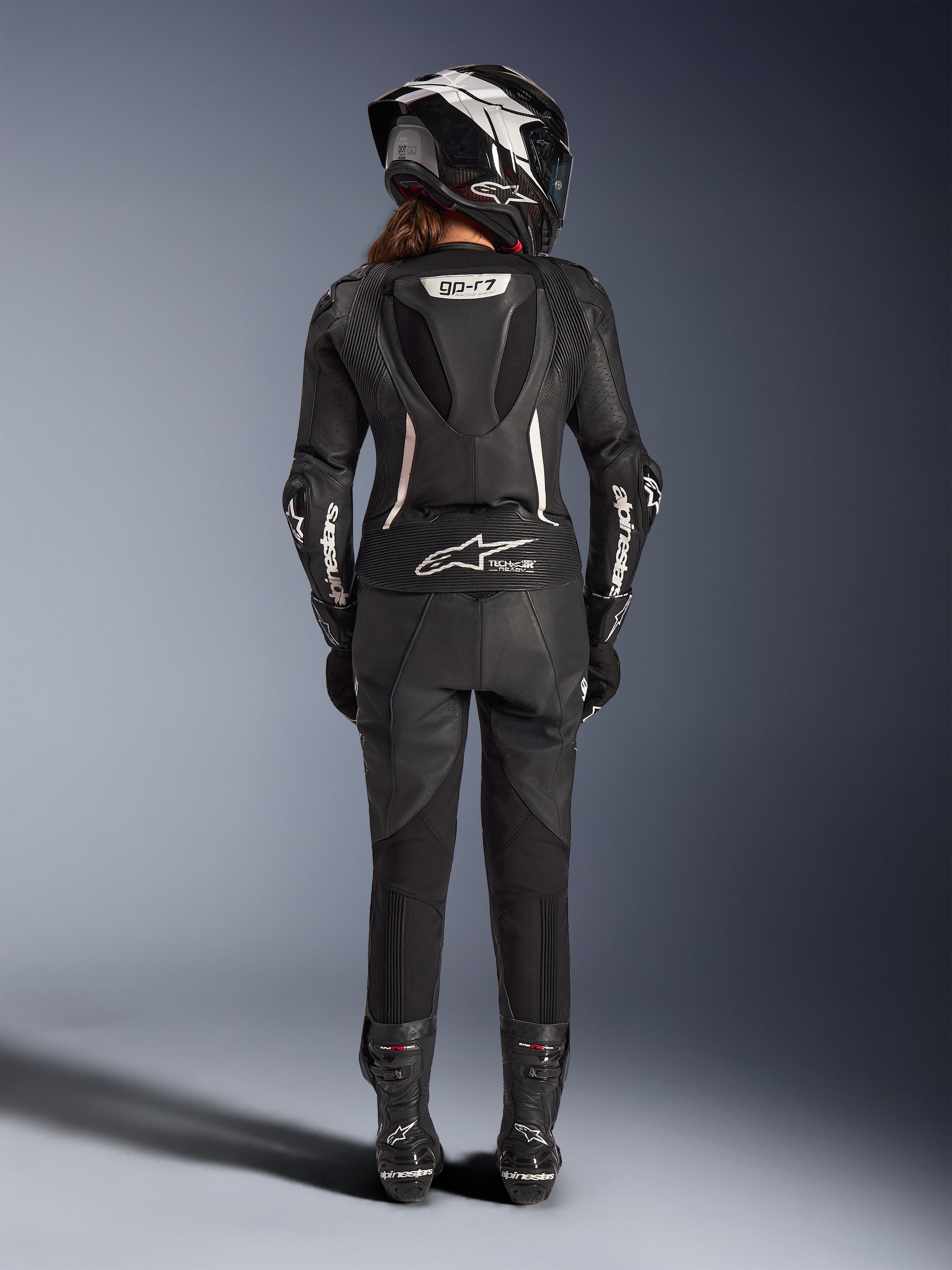 Alpinestars Stella GP-R7 2-delt læderdragt til kvinder, sort med hvide detaljer, set bagfra som viser Supertech R10 aero-pukkel og Tech-Air Ready-logo, båret med matchende full-face hjelm, racerhandsker og læderstøvler til bane- og sportskørsel
