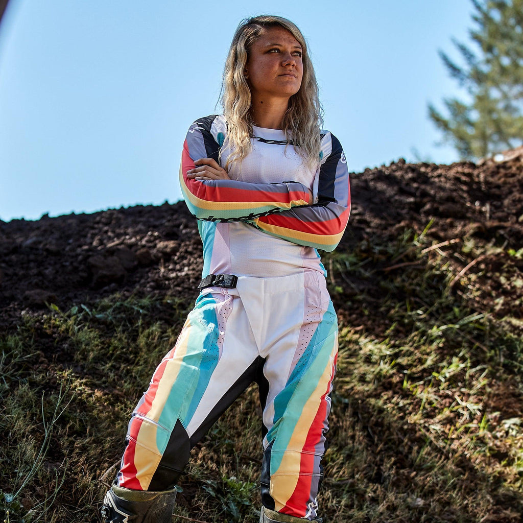 Kvindelig motocrosskører iført Alpinestars Stella 2024 Techstar-bukser og matchende jersey, Black Multicolor-design med hvid base og blå, gule og røde striber, stående med korslagte arme på en jordbane, professionelt off-road raceroutfit