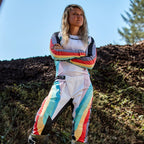 Kvindelig motocrosskører iført Alpinestars Stella 2024 Techstar-bukser og matchende jersey, Black Multicolor-design med hvid base og blå, gule og røde striber, stående med korslagte arme på en jordbane, professionelt off-road raceroutfit