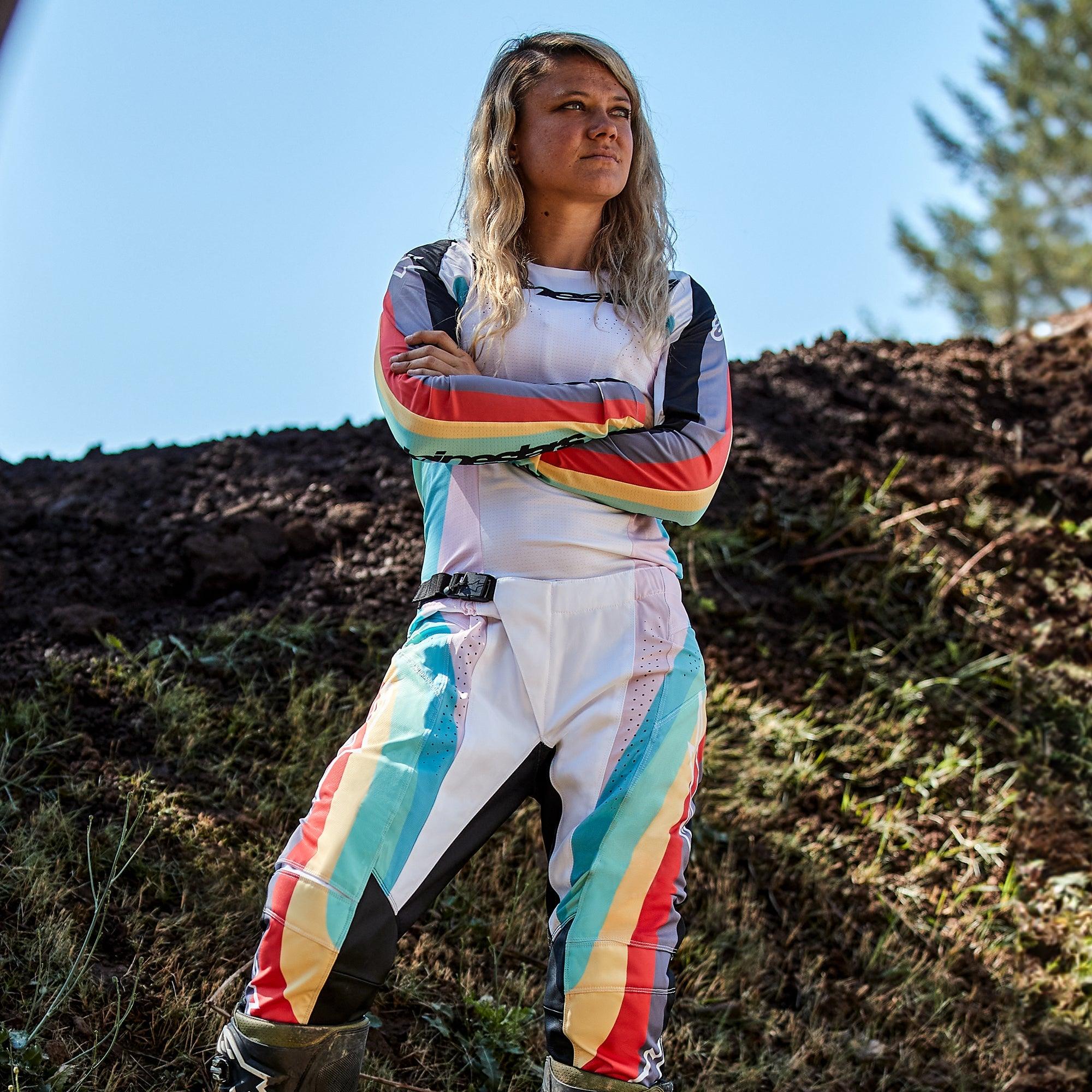 Kvindelig motocrosskører iført Alpinestars Stella 2024 Techstar-bukser og matchende jersey, Black Multicolor-design med hvid base og blå, gule og røde striber, stående med korslagte arme på en jordbane, professionelt off-road raceroutfit