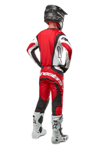 Alpinestars 2024 Techstar Ocuri bukser, motocrossbukser, Mars rød, hvid og sort, set bagfra af en motocrosskører iført fuldt racerudstyr, herunder matchende trøje, sort hjelm og hvide og sorte støvler, hvilket fremhæver Alpinestars-branding og den slidstærke tekstilkonstruktion.