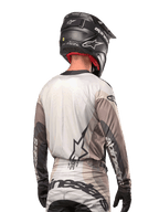 Motocross-rytter set bagfra iført Alpinestars 2024 Techstar Pneuma Jersey i Dark Sand Iron Dust Gray, kombineret med en sort hjelm med carbon-finish, med et stort stjernelogo på ryggen og Alpinestars-branding på ærmerne.