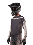 Alpinestars 2024 Techstar Pneuma trøje, motocross-racingtrøje, Dark Sand og Iron Dust Gray, langærmet performance-top med laserskårne perforeringer og hvid branding, båret med matchende bukser og sort hjelm, atletisk pasform til off-road kørsel.